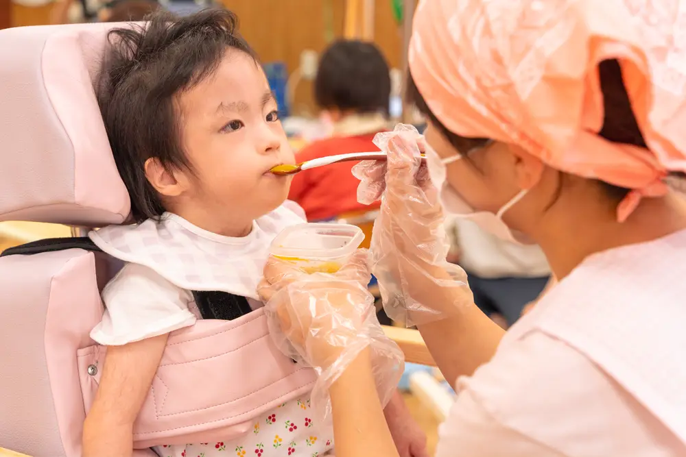 座位保持椅子で安定した姿勢で座っているお子さんが、スタッフのサポートを受けながら昼食を食べている様子。