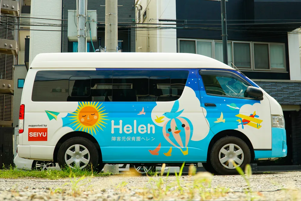 青を基調に、空に浮かぶ太陽や気球をモチーフにした「障害児保育園ヘレン」のロゴが描かれた送迎バス。