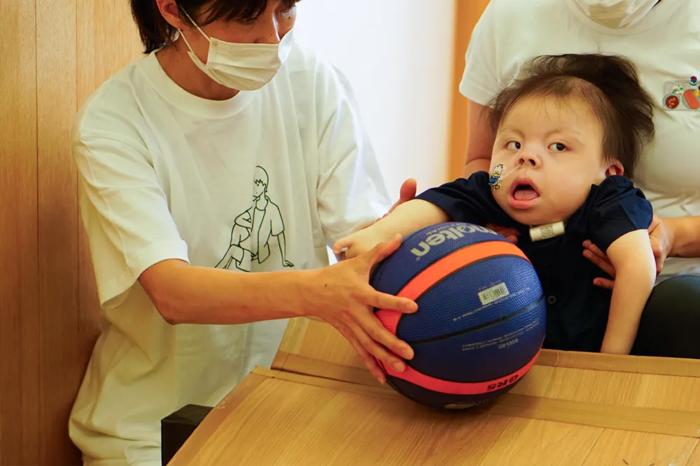 スタッフに支えられ、気管切開と経鼻チューブを付けたお子さんが、斜面台からボールを転がしている様子。
