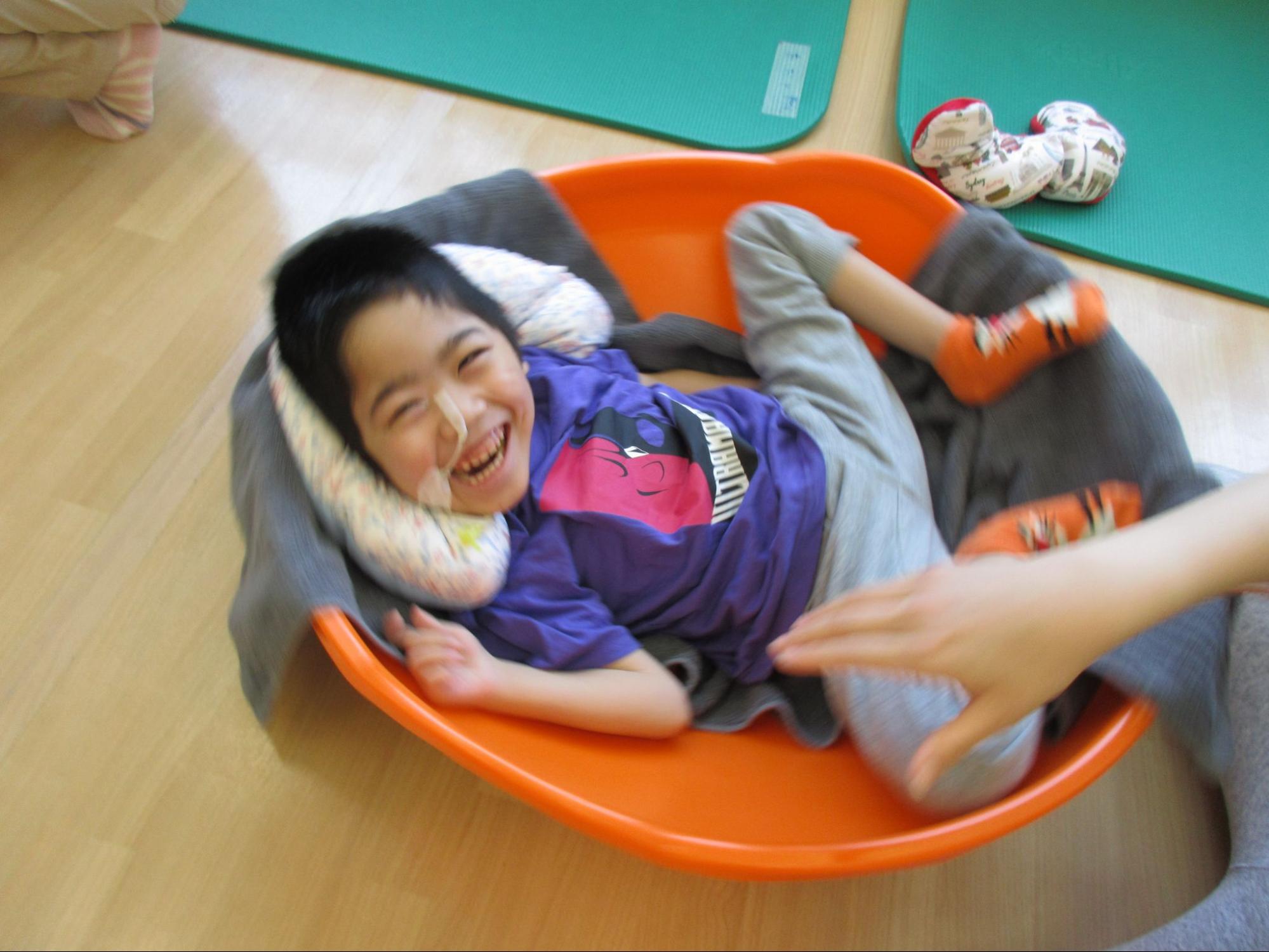 男の子が丸い大きなお椀のような形をした器具の中に横たわり、ぐるぐる回っている。楽しさで興奮してはちきれそうな笑顔でいる。